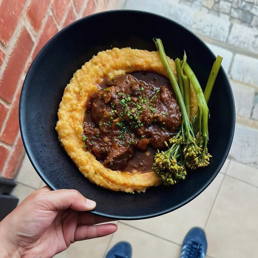 Prato preto com carne estufada em molho escuro sobre puré de batata-doce, com broccolini ao lado