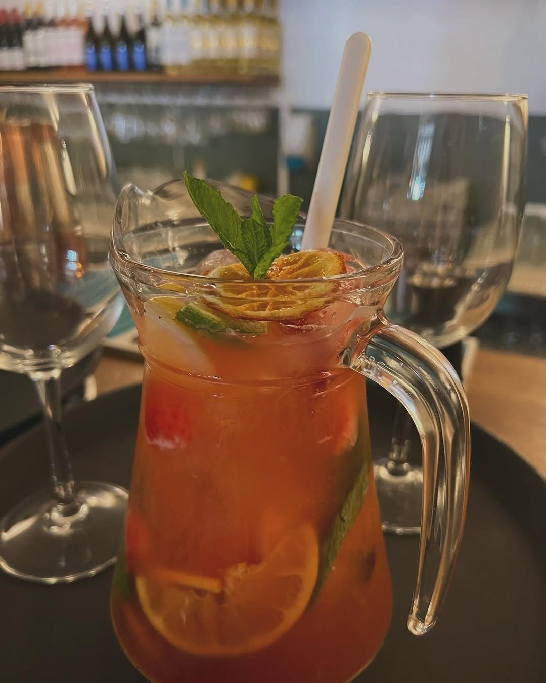 Jarro de sangria com laranja, morango e hortelã, entre dois copos de vinho, com garrafeira ao fundo