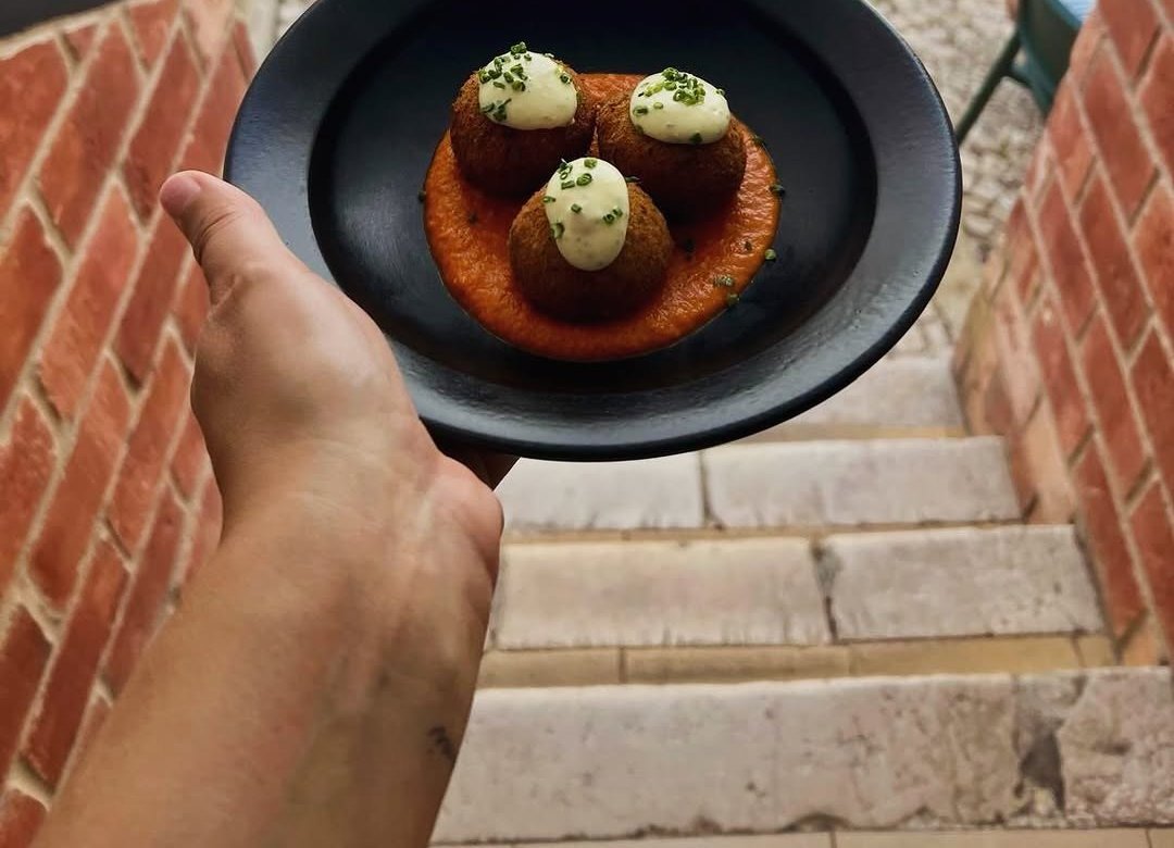 Prato preto com três bolinhos panados sobre molho laranja, segurado à mão sobre uma escada de pedra